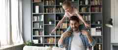 Gothaer Hausratversicherung: Vater hat Tochter auf seinen Schultern. Sie lachen beide und laufen durch ihr Wohnzimmer.